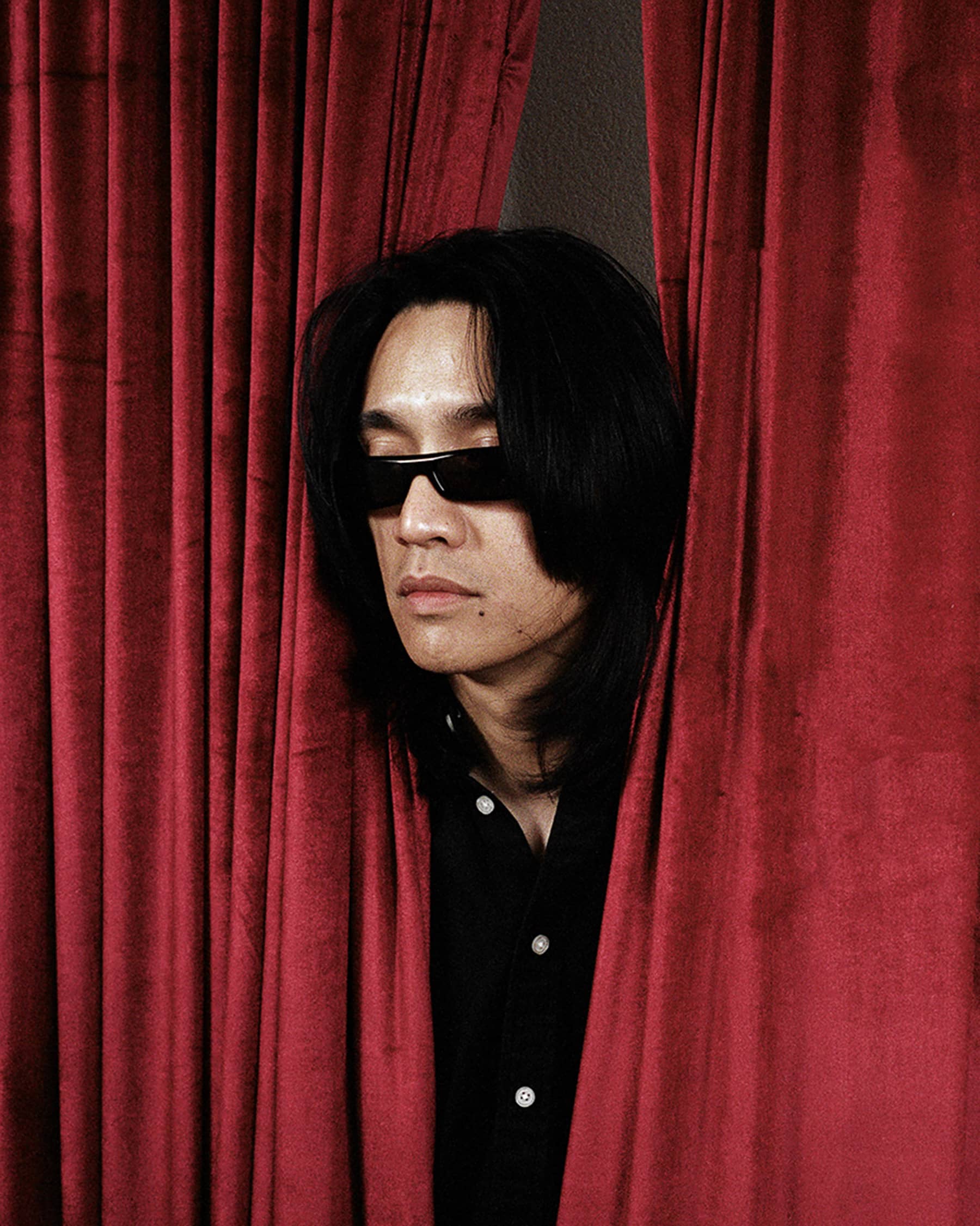 Person with sunglasses peeking through red velvet curtains