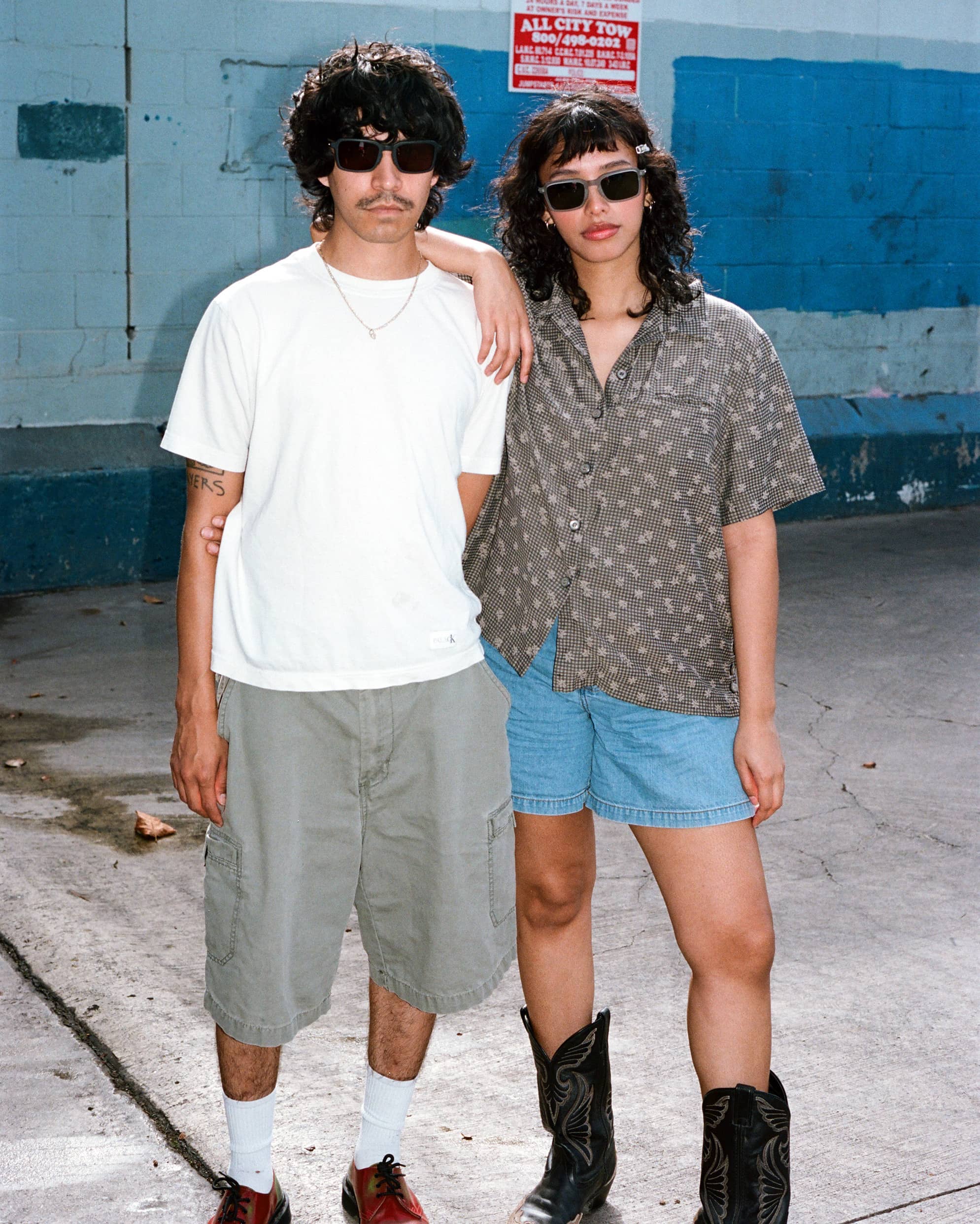 Two people in sunglasses posing against a wall, with one arm around the other's shoulder